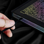decorative black journal spine with holographic foiling sits on a background of black draped silk. Hand is holding a bookmark ribbon coming out of journal