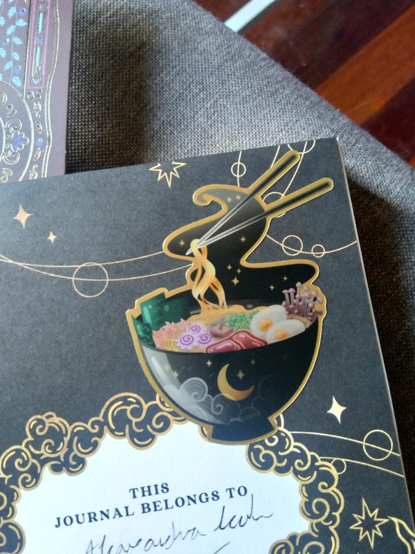 One of The Quirky Cup Collection vinyl stickers in use. A mystical black ramen sticker is stuck to the inside of a black Quirky Cup Collective journal.