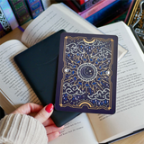 Image shows a hand gently touching a kindle and a kindle sticker insert sitting on top of the device. They sit on a bed of open books. The navy, blue, gold and white design shows a moon in the centre, surrounded by sun rays, clouds, stars, raindrops and decorative celestial embellishments.