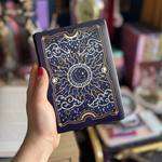 Image shows a hand holding a kindle showing off a kindle sticker insert that is placed on the back of the device. The navy, blue, gold  and white design shows a moon in the centre, surrounded by sun rays, clouds, stars, raindrops and decorative celestial embellishments.