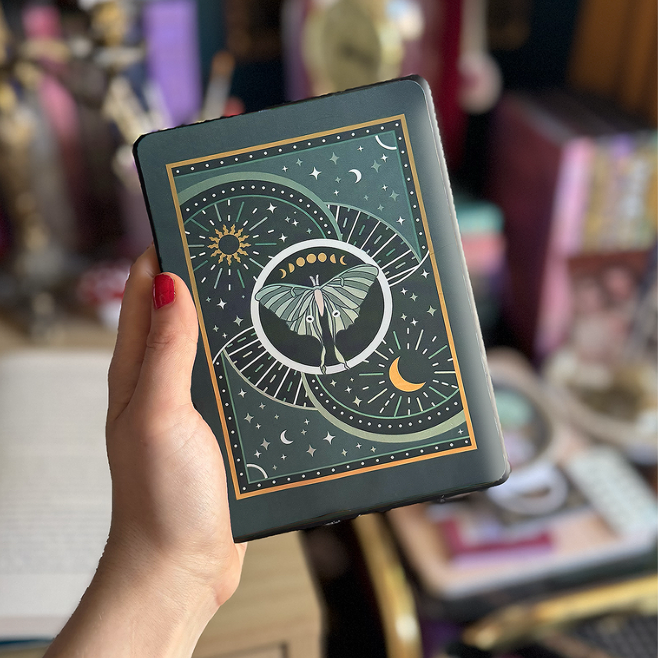 Image shows a hand holding a kindle showing off a kindle sticker insert that is placed on the back of the device. The teal, gold and white design shows a luna moth surrounded by moons, stars and decorative embellishments.