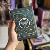 Image shows a hand holding a kindle showing off a kindle sticker insert that is placed on the back of the device. The teal, gold and white design shows a luna moth surrounded by moons, stars and decorative embellishments.