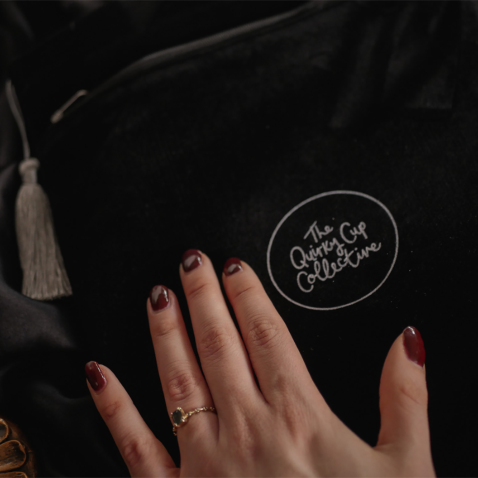 A close-up of a hand gently resting on a black velvet book sleeve embossed with "The Quirky Cup Collective" logo in white. The sleeve features a luxurious texture, paired with a silver tassel zipper pull.