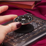 Hand holding a round, decorative clingie MagSafe device grip with intricate designs against a black fabric background.