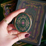 The green Literati clingie by The Quirky Cup Collective is held in front of a matching kindle sticker insert and e-reader sleeve. The oval shaped gold kindle grip features an intricate classic-style design with pink, green and gold detailing.