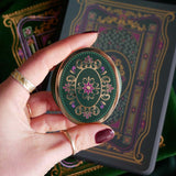 The green Literati clingie by The Quirky Cup Collective is held in front of a matching kindle sticker insert and e-reader sleeve. The oval shaped gold kindle grip features an intricate classic-style design with pink, green and gold detailing.