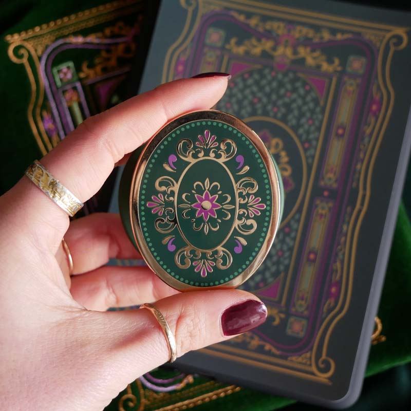 The green Literati clingie by The Quirky Cup Collective is held in front of a matching kindle sticker insert and e-reader sleeve. The oval shaped gold kindle grip features an intricate classic-style design with pink, green and gold detailing.