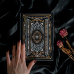 A hand holding the Black Literati Journal by The Quirky Cup Collective, showcasing its intricate gold and white floral and geometric cover design on a black background. The journal is positioned on top of a dark, silky fabric, with dried pink roses placed next to it, creating a moody, vintage-inspired scene. The journal's ribbon bookmark is partially visible, adding a touch of elegance to the overall aesthetic.