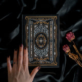 A hand holding the Black Literati Journal by The Quirky Cup Collective, showcasing its intricate gold and white floral and geometric cover design on a black background. The journal is positioned on top of a dark, silky fabric, with dried pink roses placed next to it, creating a moody, vintage-inspired scene. The journal's ribbon bookmark is partially visible, adding a touch of elegance to the overall aesthetic.