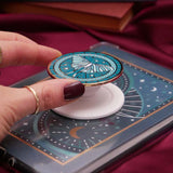 fingers holding a decorative kindle grip with intricate green and teal designs with gold accents and a luna moth on a dark fabric background