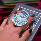 Hand holding a decorative kindle grip with intricate green and teal designs with gold accents and a luna moth on a dark fabric background