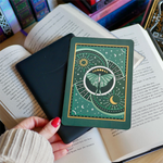 Image shows a hand gently touching a kindle and a kindle sticker insert sitting on top of the device. They sit on a bed of open books. The teal, gold and white design shows a luna moth surrounded by moons, stars and decorative embellishments.