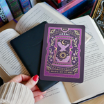 Image shows a hand gently touching a kindle and a kindle sticker insert sitting on top of the device. They sit on a bed of open books. The tarot inspired purple designs shows an hourglass with clouds, stars sun and a moon surrounded by a floral wreath surrounded by moons, stars and celestial decorative embellishments.