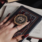 The Magic Weaver clingie kindle grip by The Quirky Cup Collective is held by . A hand holds the kindle in front of a chess board on a black table covered in books. The romantasy inspired products are all black with red, white and gold detailing and all feature motifs of a swords, crowns, snakes and hands with swiling magic. hand with long red nails over a decorated kindle on top of a book. The e-reader grip shows two hands swirling with magic with an ornate border in black, gold and white tones.