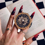The Magic Weaver clingie kindle grip by The Quirky Cup Collective is held by a hand with long red nails over a decorated kindle on top of a book. The e-reader grip shows two hands swirling with magic with an ornate border in black, gold and white tones.