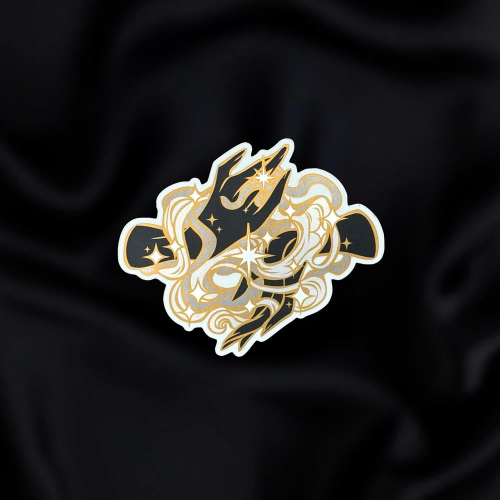 Magic Weaver- a fantasy vinyl sticker by The Quirky Cup Collective as part of the 'Serpents and Swords' collection- sits on a black fabric background. The fantasy book sticker shows two hands actively swirling gold , silver and white whisp-style magic, stars surrounding it.