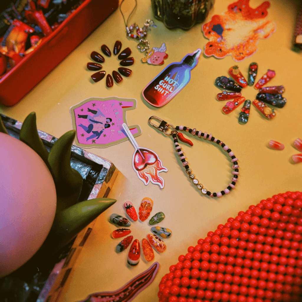 Image of The Quirky Cup Collective stickers in a styled photo with colourful fake nails, a beaded phone holder, a plant and bag.
