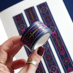 A close up of the Oath breaker washi tape by The Quirky Cup Collective. The roll of fantasy inspired washi tape is held by a hand in front of a journal showcasing the fantasy inspired stationery. The romantasy washi tape design is blue and pink with gold detailing- the intricate design showing a sword alongside geometric patterns.