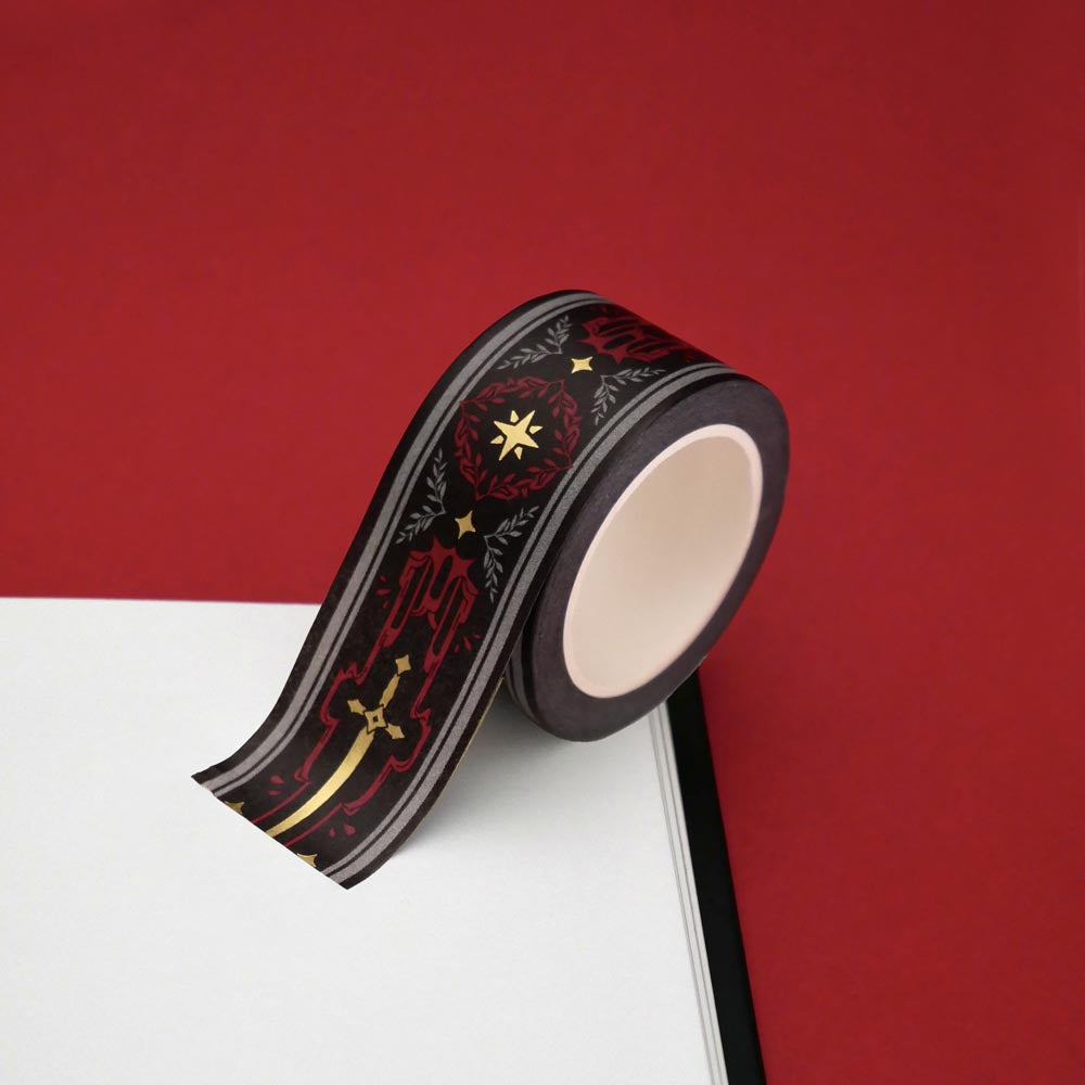 A close up of the Oath breaker washi tape by The Quirky Cup Collective. The roll of fantasy inspired washi tape is on the corner of a white journal on a red background. The romantasy washi tape design is red and black with gold detailing- the intricate design showing a sword alongside geometric patterns.