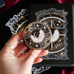 The black Otherworldly clingie by The Quirky Cup Collective is held in front of a matching kindle sticker insert and e-reader sleeve. The gold kindle grip features an intricate celestial inspired design of moons and stars in a minimal silver, gold, black and white colour palette.