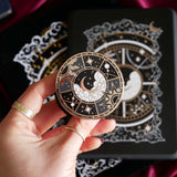 The black Otherworldly clingie by The Quirky Cup Collective is held in front of a matching kindle sticker insert and e-reader sleeve. The gold kindle grip features an intricate celestial inspired design of moons and stars in a minimal silver, gold, black and white colour palette.