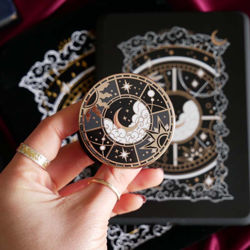 The black Otherworldly clingie by The Quirky Cup Collective is held in front of a matching kindle sticker insert and e-reader sleeve. The gold kindle grip features an intricate celestial inspired design of moons and stars in a minimal silver, gold, black and white colour palette.