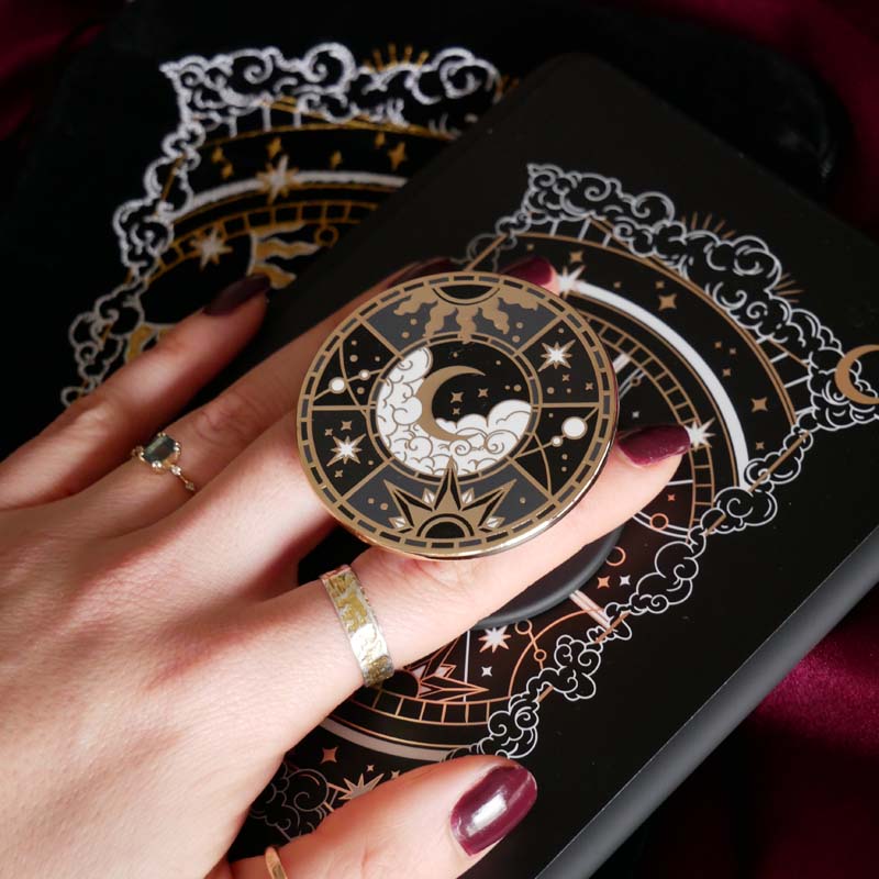 The black Otherworldly clingie by The Quirky Cup Collective is held in front of a matching kindle sticker insert and e-reader sleeve. The gold kindle grip features an intricate celestial inspired design of moons and stars in a minimal silver, gold, black and white colour palette.