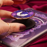 A detail shot of Fingers holding a decorative kindle grip with intricate purple designs with gold accents on a dark fabric background
