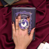 Hand holding a decorative kindle grip with intricate purple and gold designs with gold accents and a luna moth on a dark fabric background and dark nails
