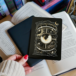 Image shows a hand gently touching a kindle and a kindle sticker insert sitting on top of the device. They sit on a bed of open books. The black, gold and white design shows a moon in the centre, surrounded by a sun, clouds, stars and decorative celestial embellishments.