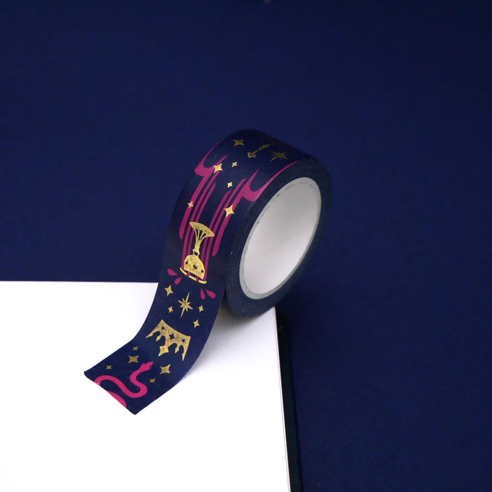 A close up of the Blood Spilled washi tape by The Quirky Cup Collective. The roll of fantasy inspired washi tape is on the corner of a white journal on a blue background. The fantasy book inspired washi tape design is pink, blue with gold detailing with a goblet motif.