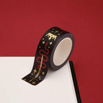 A close up of the Prophecy Foretold washi tape by The Quirky Cup Collective. The roll of fantasy inspired washi tape is on the corner of a white journal on a red background. The bookish washi tape design is black with red and gold detailing- the intricate design showing a crown, red snake, goblet and star icons.