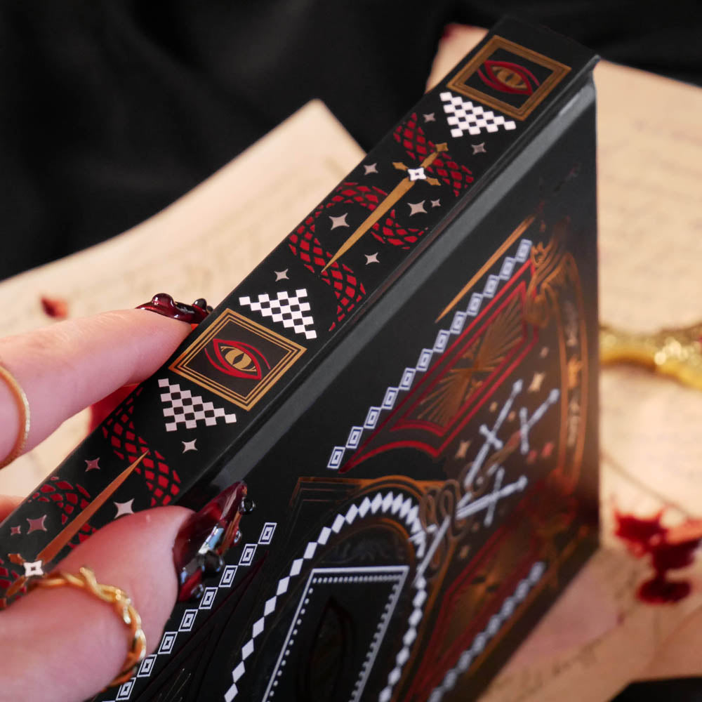 A close up shot of the 'Destiny Awaits' reading companion journal by The Quirky Cup Collective. On a background of handwritten notes, the spine of the black book is shown with the intricate red, gold and red foil showcasing the intricate designs of a dragon eye, swords, stars and intricate geometric patterns.