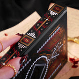 A close up shot of the 'Destiny Awaits' reading companion journal by The Quirky Cup Collective. On a background of handwritten notes, the spine of the black book is shown with the intricate red, gold and red foil showcasing the intricate designs of a dragon eye, swords, stars and intricate geometric patterns.