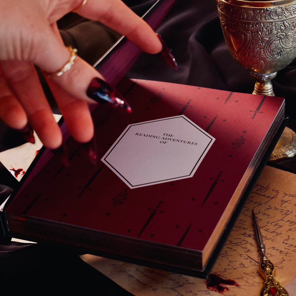 In a dark scene, the front page of a Quirky Cup Collective reading journal is held open to show a nameplate on a lush red spread with snake and sword motifs. The journal is help open by a hand with long red nails in a scene with a goblet and several handwritten notes.