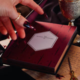 In a dark scene, the front page of a Quirky Cup Collective reading journal is held open to show a nameplate on a lush red spread with snake and sword motifs. The journal is help open by a hand with long red nails in a scene with a goblet and several handwritten notes.