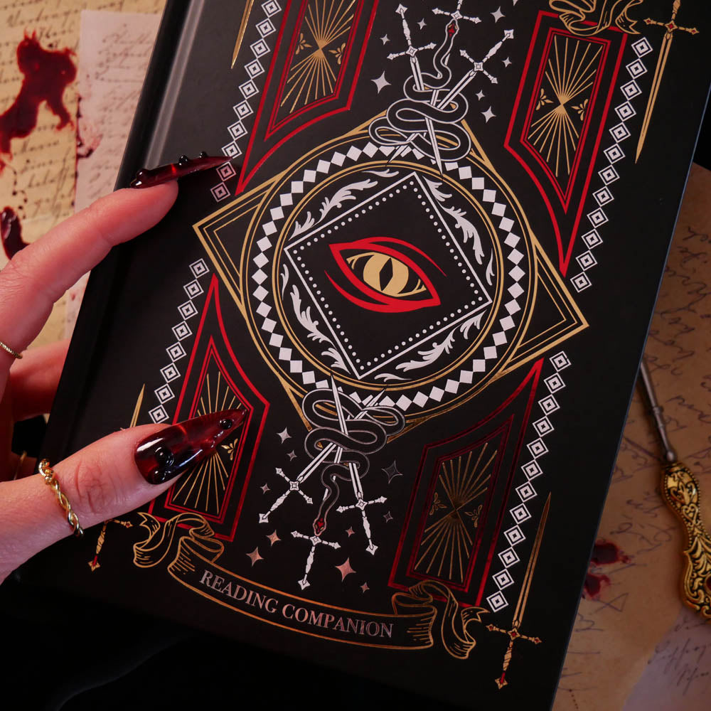 A detail shot of the Quirky Cup Collective 'Destiny Awaits' reading Companion. The black journal features a dragon eye design alongside sword, snake and crown motifs in red, gold and white foils. A hand with intricate red nails holds the journal in front of a stack of handwritten notes covered in blood.