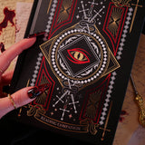 A detail shot of the Quirky Cup Collective 'Destiny Awaits' reading Companion. The black journal features a dragon eye design alongside sword, snake and crown motifs in red, gold and white foils. A hand with intricate red nails holds the journal in front of a stack of handwritten notes covered in blood.