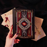 On a dark background sits the Quirky Cup Collective Destiny Awaits reading Companion. The black journal features a dragon eye design alongside sword, snake and crown motifs in red, gold and white foils. A hand with intricate red nails holds the journal in front of a stack of handwritten notes covered in blood.