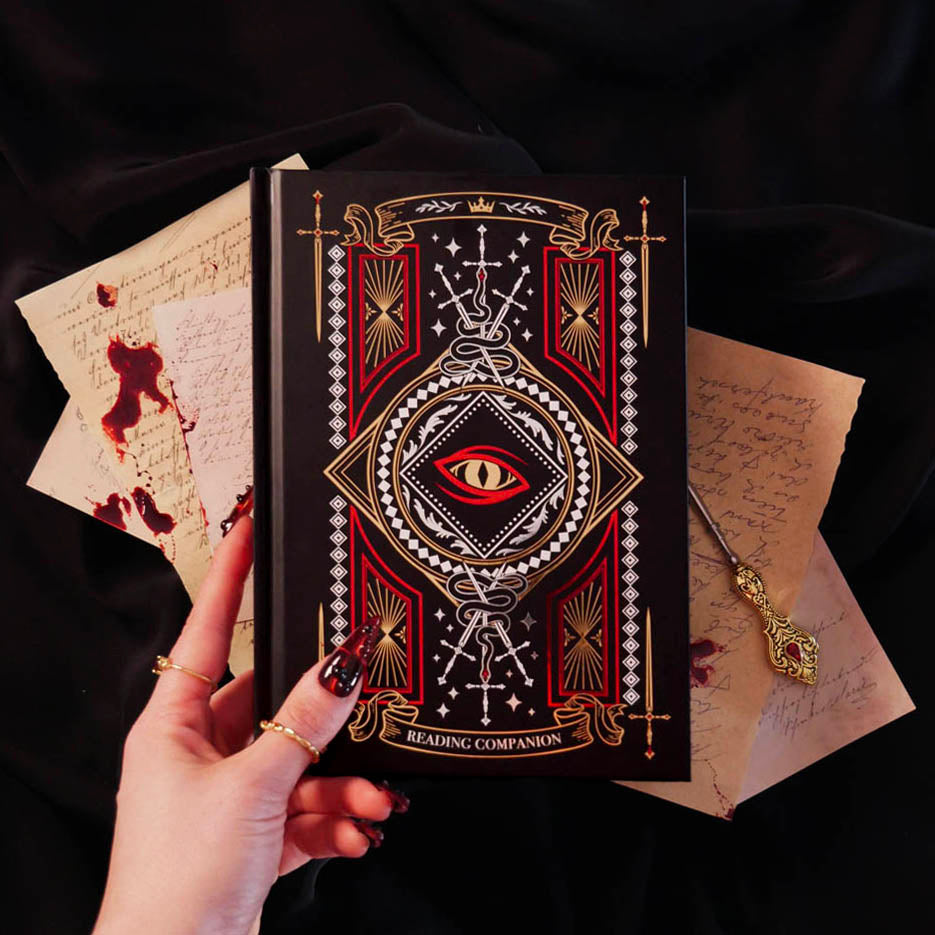 On a dark background sits the Quirky Cup Collective Destiny Awaits reading Companion. The black journal features a dragon eye design alongside sword, snake and crown motifs in red, gold and white foils. A hand with intricate red nails holds the journal in front of a stack of handwritten notes covered in blood.