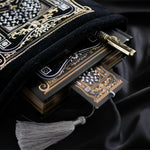 A hand holding a luxurious reading journal with an intricate gold and white design, accompanied by an ornate matching pen. The journal lies on top of a black velvet book sleeve from The Quirky Cup Collective, showcasing a coordinated design. A matching bookmark with a silver tassel is also partially visible. All items are set against a smooth black satin fabric background, creating an elegant and cohesive visual presentation.