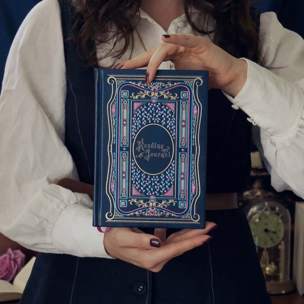 A woman holds a royal blue Reading Journal by The Quirky Cup Collective. The high end blue reading journal features intricate gold, pink and blue foiled illustrations and the woman wears a white shirt and navy pinafore.