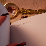 An open Serpents & Swords Reading journal by The Quirky Cup collective shows the 'favourite' reading journal spread. The journal is held open by a hand with intricate red fingernails in a dark and shadowy setting surrounded by handwritten notes and a magnifying glass.