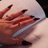 An open Serpents & Swords Reading journal by The Quirky Cup collective shows the 'bookshelf' reading journal spread. The journal is held open by a hand with intricate red fingernails in a dark and shadowy setting surrounded by handwritten notes and scrolls.