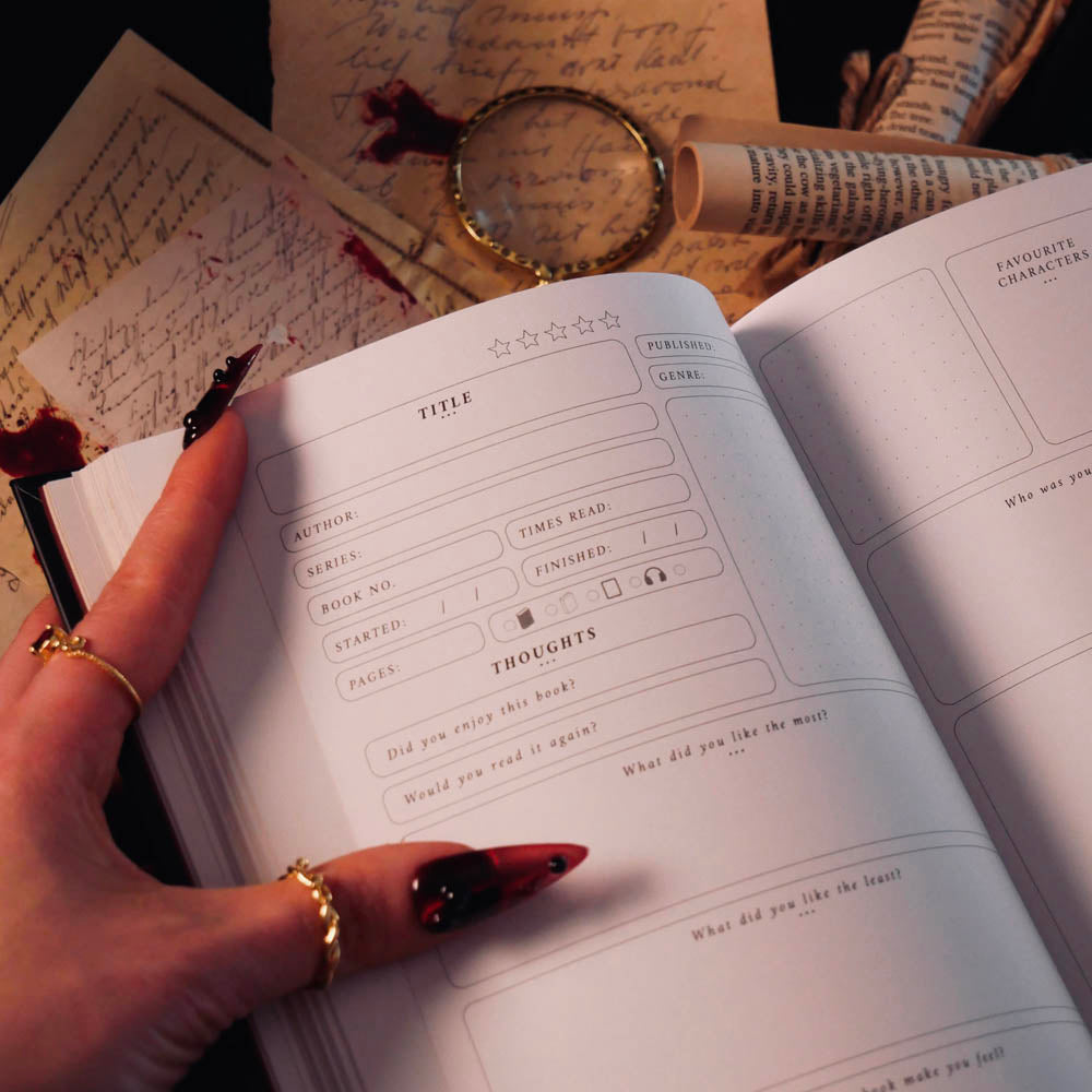 An open Serpents & Swords Reading journal by The Quirky Cup collective shows the 'book reviews' reading journal spread. The journal is held open by a hand with intricate red fingernails in a dark and shadowy setting surrounded by handwritten notes and scrolls.