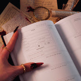 An open Serpents & Swords Reading journal by The Quirky Cup collective shows the 'book reviews' reading journal spread. The journal is held open by a hand with intricate red fingernails in a dark and shadowy setting surrounded by handwritten notes and scrolls.