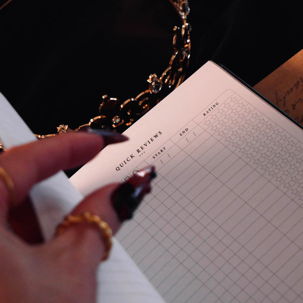 An open Serpents & Swords Reading journal by The Quirky Cup collective shows the 'quick reviews' reading journal spread. The journal is held open by a hand with intricate red fingernails in a dark and shadowy setting surrounded by handwritten notes and scrolls.