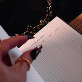 An open Serpents & Swords Reading journal by The Quirky Cup collective shows the 'quick reviews' reading journal spread. The journal is held open by a hand with intricate red fingernails in a dark and shadowy setting surrounded by handwritten notes and scrolls.