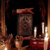 A promo image for the Serpents & Swords collection. Hands hold a lush reading companion in front of a mirror, candles and goblets surrounding the scene. The black journal features a dragon eye motif alongside swords and geometric patterns.
