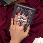 Heart with wings E-reader and phone grip held by a hand with dark nail polish against a black background